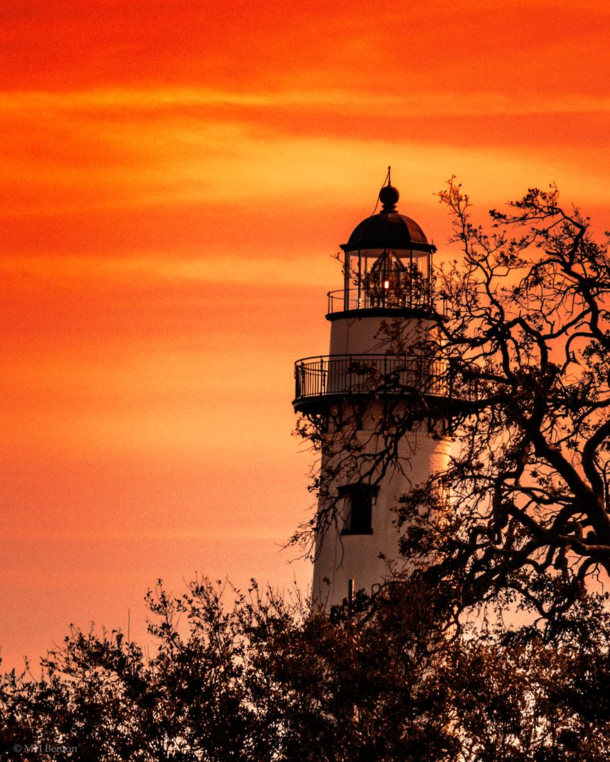 The St. Simons Island Lighthouse lantern glowing against an orange sunset sky, framed by live oak branches